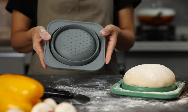 A person holding a collapsed grey KEVJES PopOut silicone pizza dough container, displaying the honeycomb release bottom.