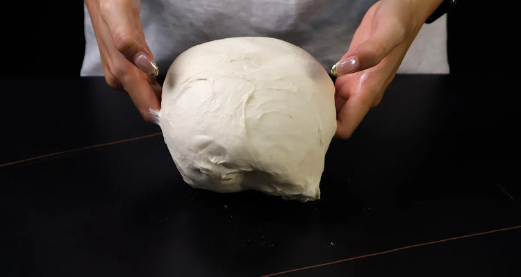 Hands lifting soft pizza dough during folding on a dark work surface