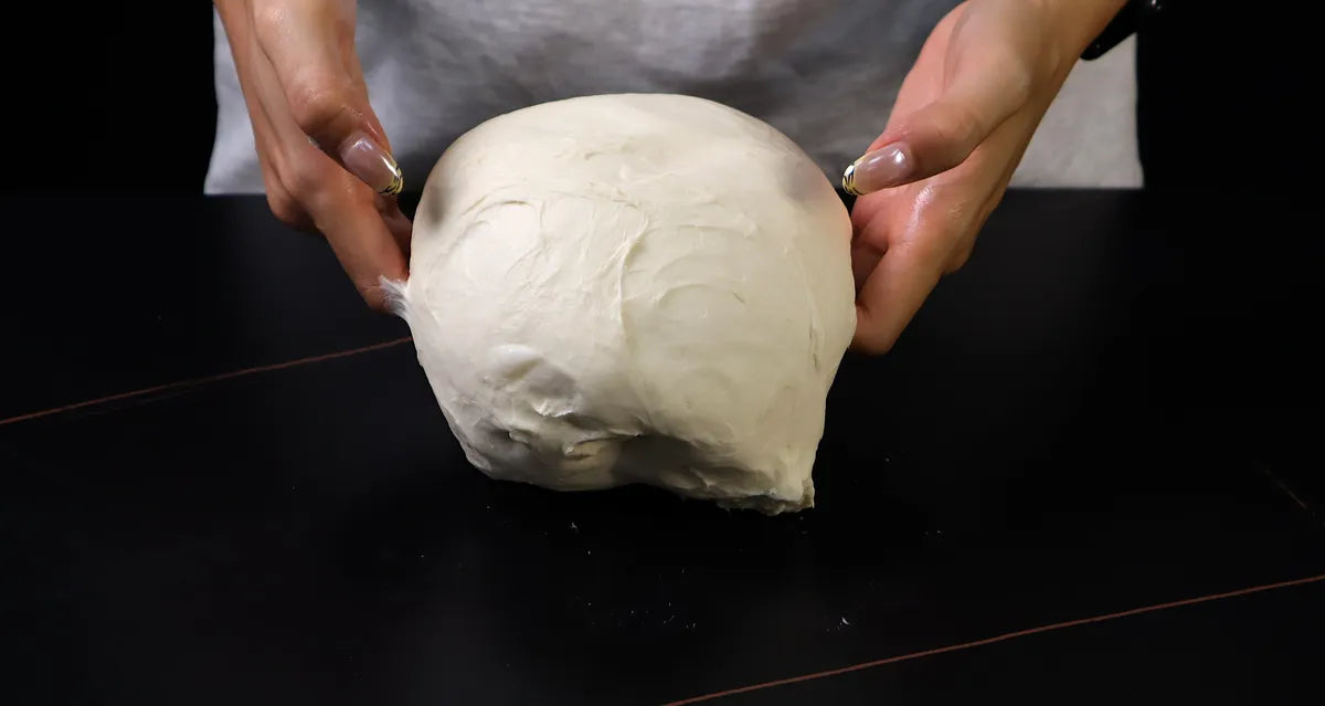 Hands lifting soft pizza dough during folding on a dark work surface